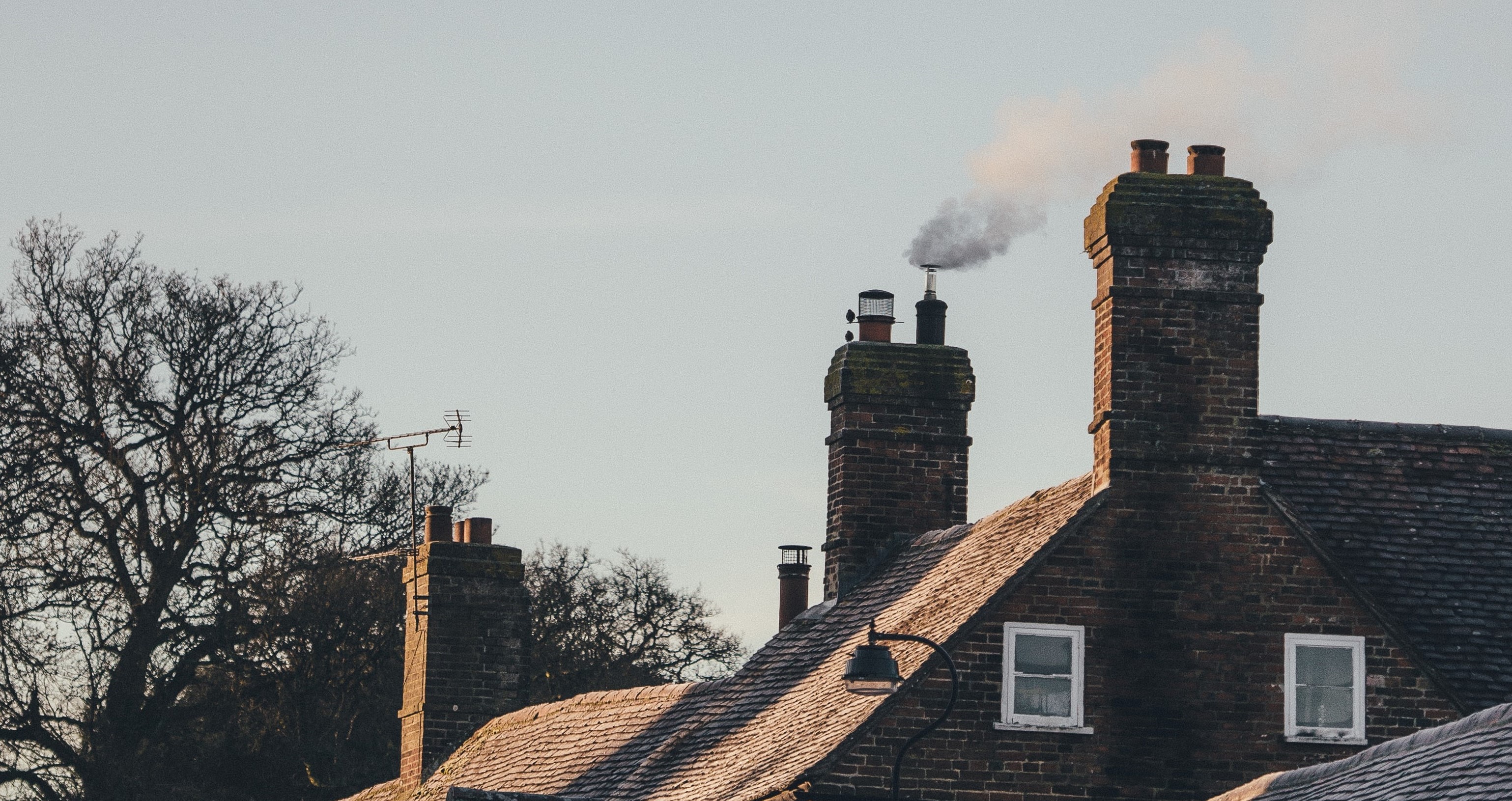 A few chimneys in a row, with smoke coming out of the one in the middle. Only the roofs of the houses can be seen, as well as some of the brick wall of the first in the row, and a grey sky. There are a few trees in the background with no leaves, so it's clearly autumn.