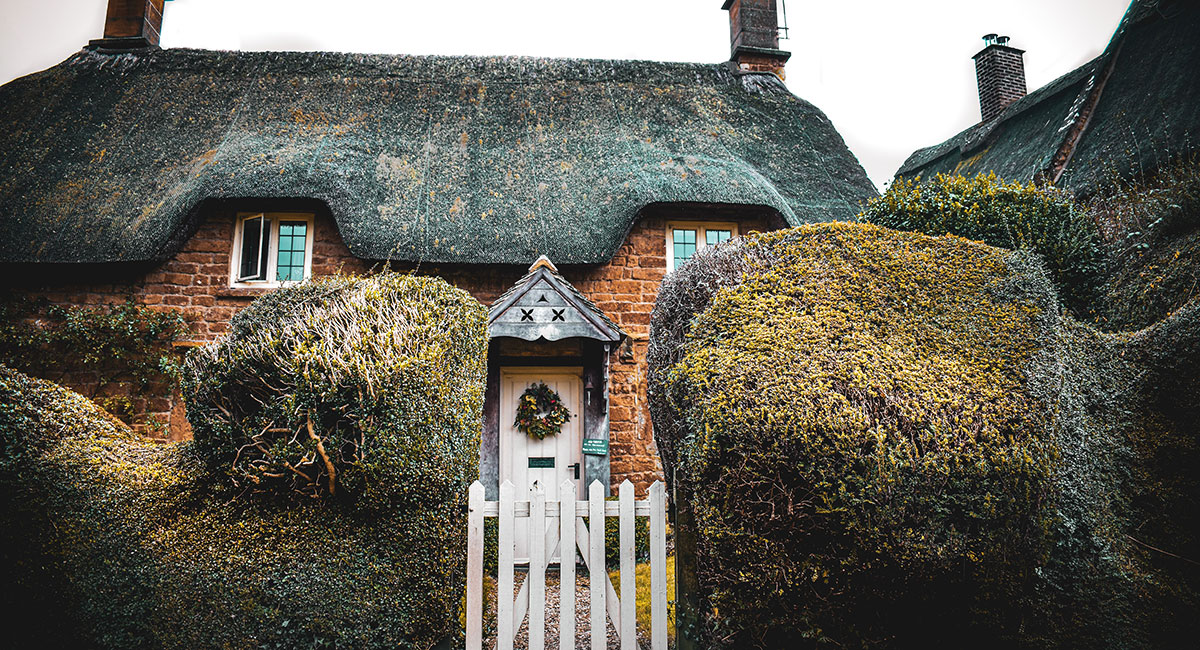 A thatched red brick cottage. There is a wreath on the white front door and a white picket fence outside. 