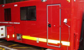 The pod is a red block, with a neon yellow stripe along the bottom. Two small windows are visible, as well as the door to get inside. Devon and Somerset Fire and Rescue Service is written in white capitals by the left window. A small storage space has its doors open underneath the window.