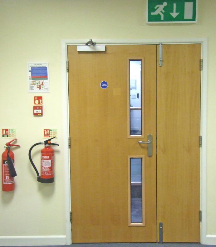 A wooden fire door on a cream wall with a green fire exit sign above it. Two fire extinguishers and fire safety signs are next to it. 
